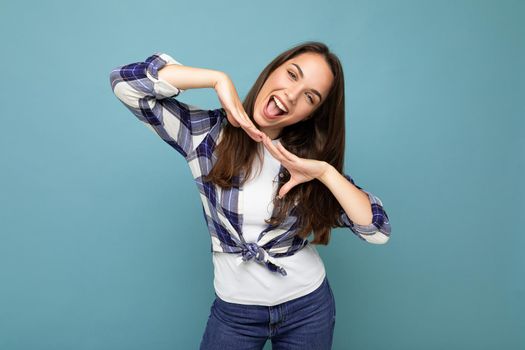 Photo shot of young positive delightful smiling pretty brunette woman with sincere emotions wearing hipster check shirt standing isolated on blue background with free space.