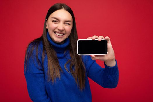 Smiling beautiful happy young brunette woman wearing casual blue sweater isolated over red background with empty space holding in hand mobile phone and showing smartphone with empty screen for mockup looking at camera.