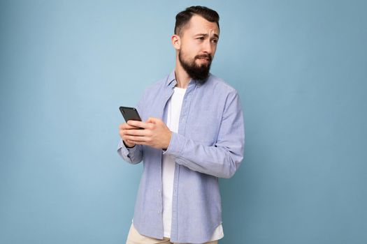 Thoughtful handsome young man isolated over background wall wearing everyday clothes holding and using mobile phone writing sms looking to the side and thinking.