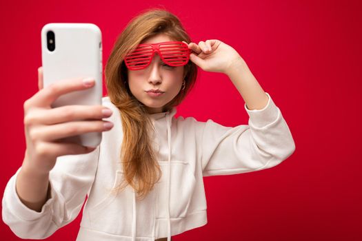 Closeup portrait of attractive positive smiling young blonde woman wearing stylish white hoodie and funny colorful glasses standing isolated over colourful background holding and using mobile phone taking selfie photo looking at gadjet.