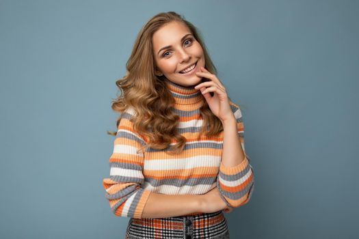Portrait of young winsome attractive happy smiling blonde woman with wavy-hair wearing striped sweater isolated over blue background with empty space.