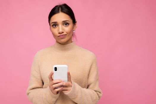 Attractive cute unhappy young brunet woman wearing beige warm sweater standing isolated over pink background surfing on the internet via phone looking at camera.