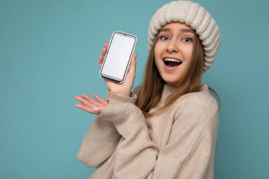 Closeup Photo of amazed beautiful positive good looking young woman wearing stylish beige sweater and beige knitted winter hat poising isolated over blue background with empty space holding in hand and showing mobile phone with empty screen for mockup looking at camera.