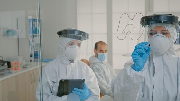 Team of stomatologists wearing ppe suits using tablet and black marker. Dental specialist drawing tooth model for reference on stomatology cabinet glass. Orthodontists operating during pandemic