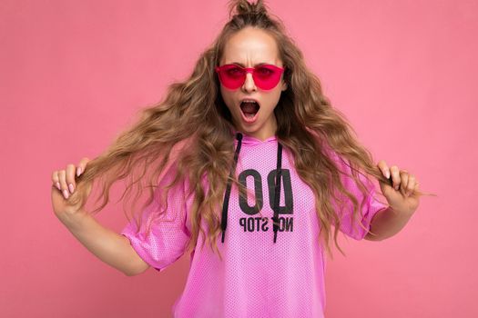 Pretty surprised positive young blonde curly woman isolated over pink background wall wearing casual pink t-shirt and stylish colourful sunglasses looking at camera and shouting.