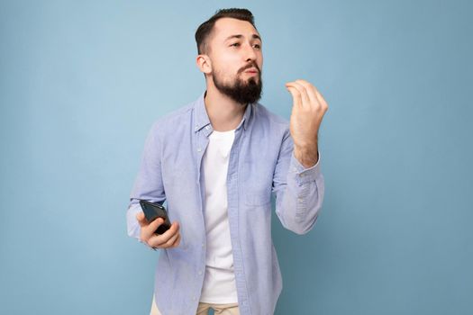 positive emotional good looking young brunet bearded man wearing casual blue shirt and white t-shirt poising isolated on blue background with empty space holding in hand mobile phone looking to the side.
