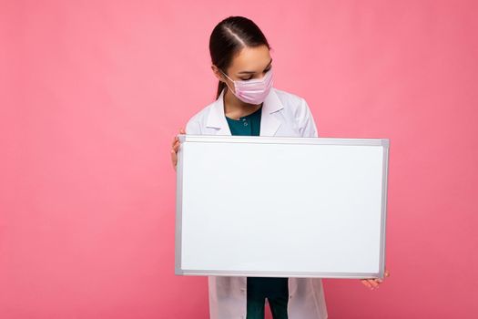 Photo of young attractive nurse in protective face mask holding an empty magnetic board with copy space for information isolated on pink background.