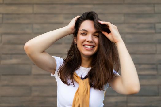 Portrait of successful smiling joyful happy young brunet woman wearing casual white t-shirt with yellow sweater poising near brown wall in the street and having fun.