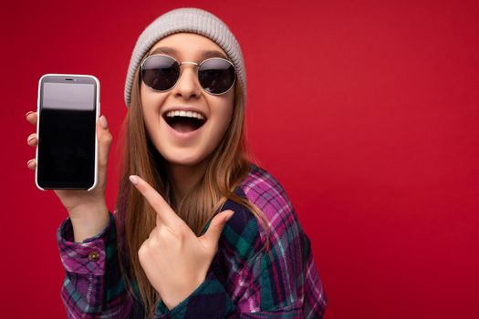 Closeup photo of attractive smiling positive young blonde woman wearing stylish purple shirt and casual white t-shirt grey hat and sunglasses isolated over red background holding in hand and showing mobile phone with empty screen for mockup looking at camera and pointing finger at gadjet display. Wow. Copy space