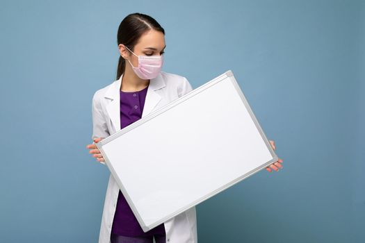 Nurse in protective face mask and white medical coat holding an empty magnetic board isolated on blue background.