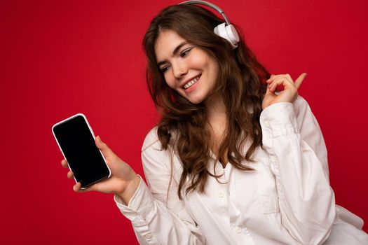 Attractive positive young brunette curly woman wearing white shirt isolated on red background wall holding and showing mobile phone with empty display wearing white bluetooth headphones listening to music looking at gadjet.