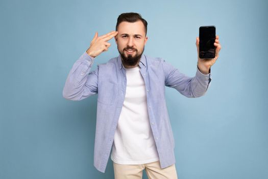 Dissatisfied handsome good looking young brunette unshaven man with beard wearing casual white t-shirt and blue shirt poising isolated on blue background with empty space holding in hand mobile with empty screen looking at camera and showing gun gesture near head.