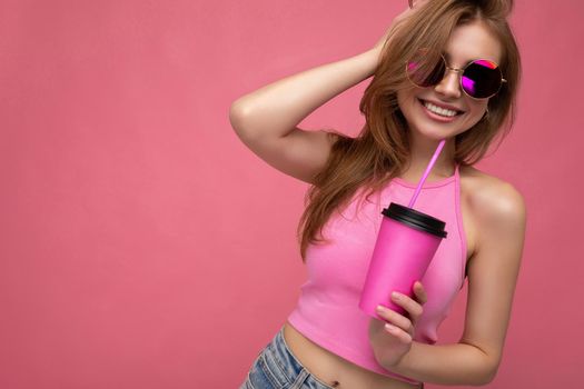Charming young happy smiling blonde woman wearing stylish pink clothes isolated over colourful background wall holding paper cup for mockup drinking milkshake looking at camera. copy space