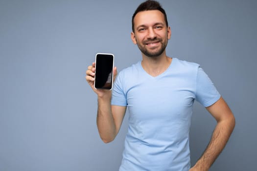 Handsome happy cool young man good looking wearing casual stylish clothes standing isolated over colourful background wall holding smartphone and showing phone with empty screen display looking at camera.