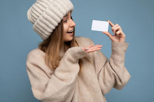 Portrait Beautiful surprised happy smiling young dark blonde female person wearing beige sweater and knitted beige hat isolated over blue background holding and showing credit card looking at plastic contactless bank card.