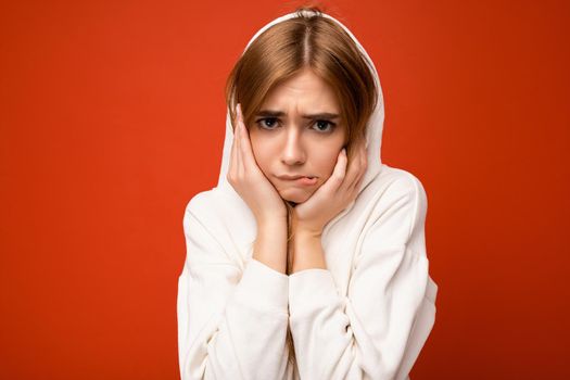 Young doubtful beautiful dark blonde woman with sincere emotions isolated on background wall with copy space wearing casual white hoodie looking at camera.