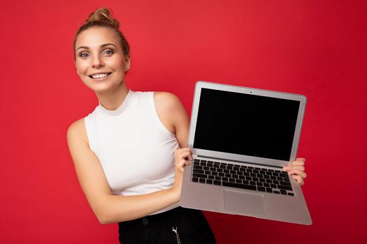 beautiful smiling fascinating happy blond young woman with gathered hair looking at camera holding computer laptop wearing white t-shirt isolated over red wall background.