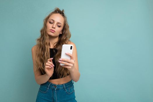 Photo shot of attractive thoughtful good looking young woman wearing casual stylish outfit poising isolated on background with empty space holding in hand and using mobile phone messaging sms looking at smartphone display screen.