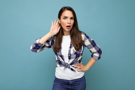 Young concentrated beautiful brunet woman with sincere emotions wearing stylish check shirt standing isolated on blue background with empty space and listening to something by putting hand on the ear.
