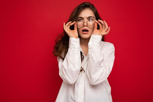 Attractive funny positive shocked amazed surprised young curly brunette woman wearing white shirt and optical glasses isolated on red background with empty space.