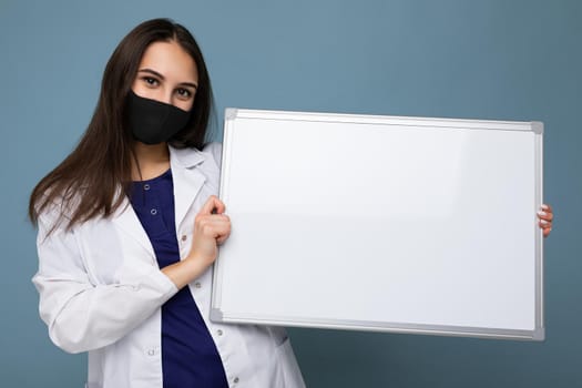 Nurse in protective face mask and white medical coat holding an empty magnetic board isolated on blue background.