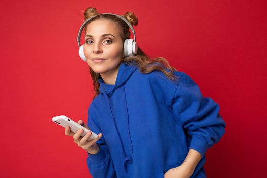 Photo of beautiful young woman wearing stylish blue hoodie isolated over red background wearing white bluetooth wireless headphones and listening to music and using mobile phone surfing on the internet looking at camera.