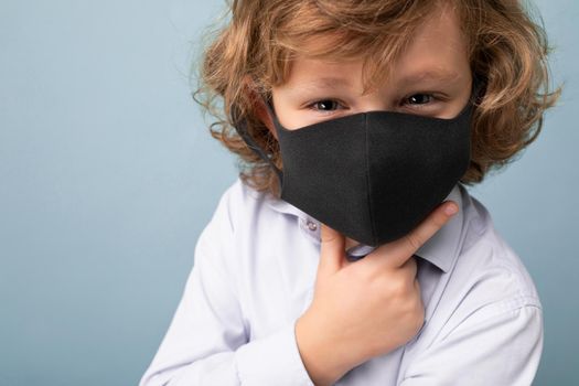 Closeup photo of handsome curly blonde little boy in medical black mask standing isolated over blue background. Coronavirus concept. Copy space