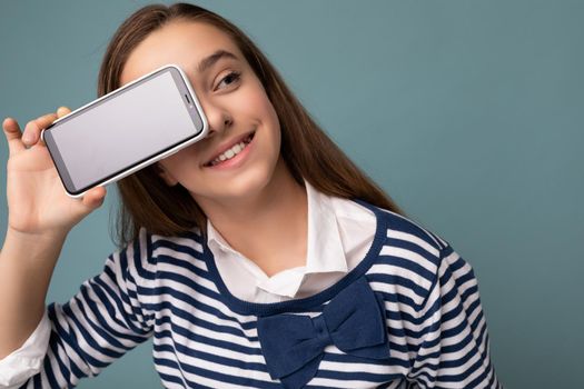 Attractive positive smiling brunette girl wearing striped longsleeve standing isolated on blue background with copy space holding smartphone showing phone in hand with empty screen display for mockup pointing at gadjet looking to the side.