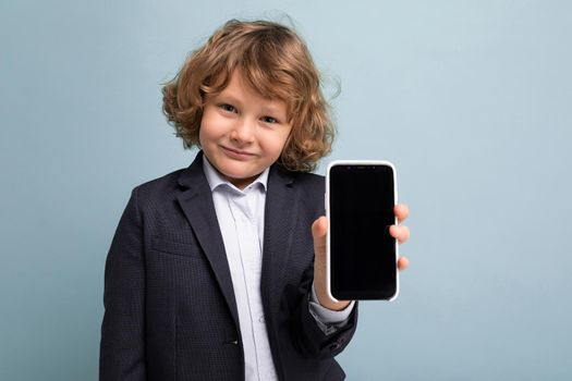 Closeup photo of Handsome positive boy with curly hair wearing suit holding phone isolated over blue background looking at camera and showing smartphone with empty display screen. Mockup
