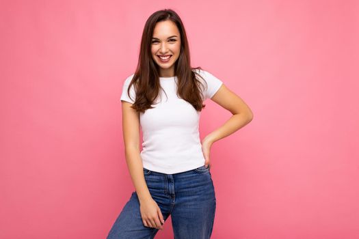 Photo portrait of young beautiful smiling hipster brunette woman in white t-shirt with mockup. Sexy carefree female person posing isolated near pink wall with empty space in studio. Positive model with natural makeup.