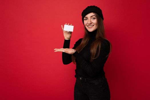 Photo of beautiful happy young brunette woman wearing black sweater and hat isolated on red background holding and showing credit card looking at camera.