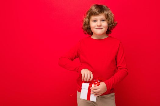 Photo of happy smiling blonde curly male kid isolated over red background wall wearing red sweater holding gift box and looking at camera. Copy space