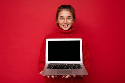 Photo of Beautiful satisfied happy young woman holding computer laptop looking at camera wearing red sweater isolated over red wall background. cutout