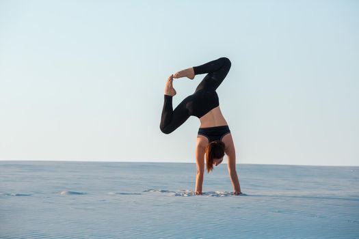 Young woman practicing inversion balancing yoga pose handstand on sand. Sunset and white sand.