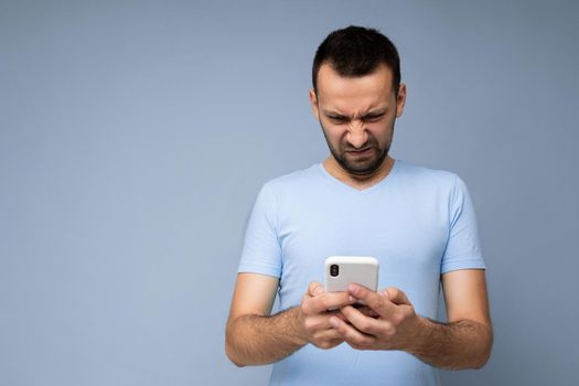 Photo of dissatisfied handsome young man with beard wearing everyday blue t-shirt isolated over blue background holding and using mobile phone communication online on the internet looking at gadjet display.
