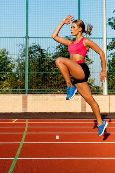 Young, beautiful girl athlete in sportswear doing warm-up at the stadium. Jumping up, one leg bent at the knee