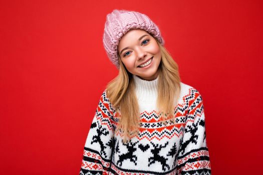 Photo of beautiful happy smiling young blonde woman isolated over red background wall wearing winter sweater and trendy pink hat looking at camera.