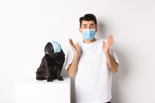 Covid-19, animals and quarantine concept. Shocked dog owner and pug wearing medical masks, staring at camera amazed, standing over white background.