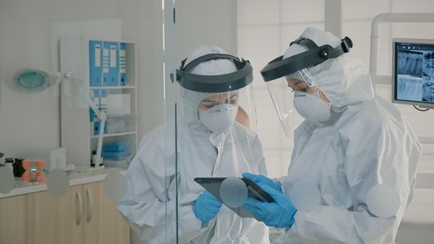 Dentistry team with virus protection suits analyzing digital tablet in dental cabinet. Stomatologists looking at device for consultation and teethcare while patient sitting on chair