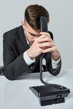 A young man in a black suit dials the phone number while sitting in the office. Manager beats himself over the head with a telephone