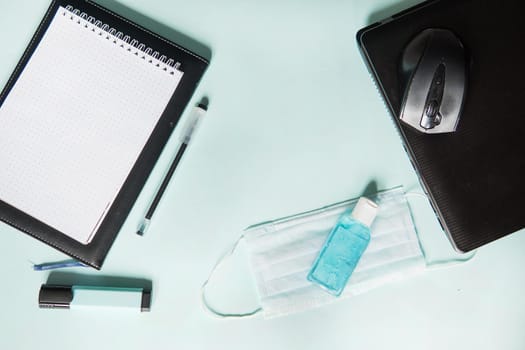 Top view of a desktop with notebooks, pens, and a laptop. Work from home using an online device, protection from coronavirus during the quarantine period. Protective masks and antiseptic on a mint background.