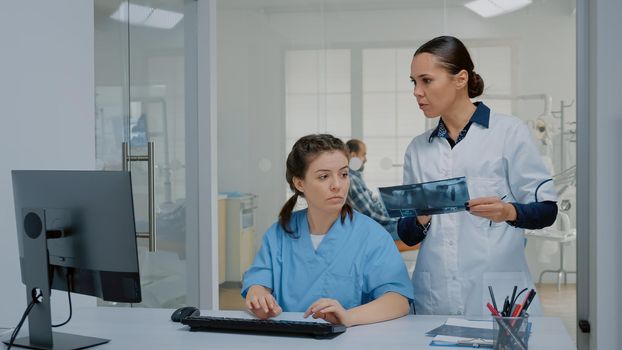 Team of nurse and stomatologist examining x ray and computer screen for professional oral care operation at dental clinic. Orthodontic staff using technology for modern radiography