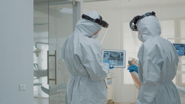 Stomatologists with ppe suits holding digital tablet with teeth x ray analyzing dental healthcare for implant operation at medical clinic. Dentists using scan technology during pandemic