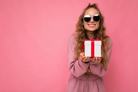 Shot of attractive positive smiling young blonde woman isolated over colourful background wall wearing everyday trendy outfit holding gift box and looking at camera. Copy space