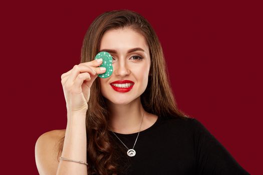 Pretty long hair woman in black dress holding chips for gambling in casino. Studio shot on red background. Poker. Two green Chips in Hand