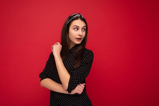 Photo of beautiful cute positive brunette girl standing isolated over red wall wearing casual stylish black clothes looking to the side and thinking.