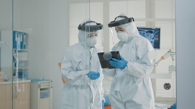 Orthodontists wearing protection suits looking at tablet for patient dental consultation at clinic. Assistant helping dentist with dentition care and modern stomatology equipment during pandemic