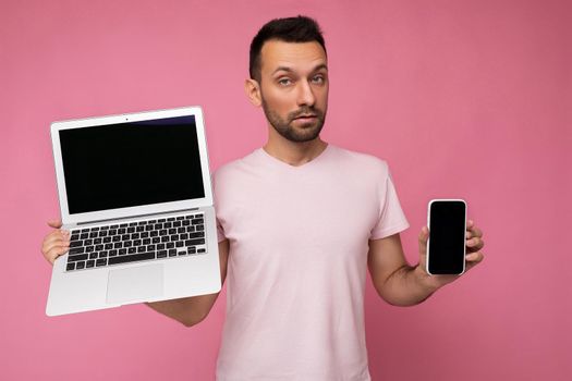 Handsome amazed brunet man holding laptop computer and mobile phone looking at camera in t-shirt on isolated pink background.