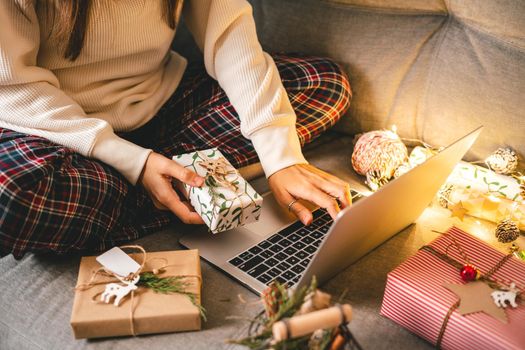 Close up of woman hands with gifts, typing at laptop. Online shopping at Christmas holidays. Cropped female sit on couch with natural eco presents and decor. Merry Christmas packing Concept.