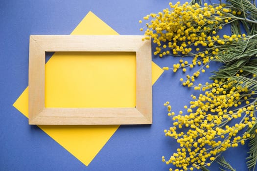 Spring concept, top view. Mimosa, a sheet of yellow paper and a gold frame on a blue background. Mimosa close-up. Happy spring. Space for text.